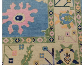 Könnte beinhalten: Nahaufnahme eines gemusterten Teppichs mit hellblauem Mittelfeld und beigem Rand. Der Teppich zeigt florale und geometrische Muster in Rosa, Grün, Gelb und Marineblau. Der Teppich hat einen Fransenrand.