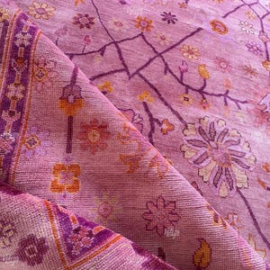May include: A folded, pink rug with a floral pattern. The rug features a repeating design of flowers and geometric shapes in shades of pink, purple, and orange. The rug's border has a detailed, geometric pattern.