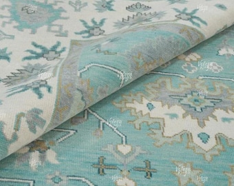 May include: A close-up of a folded area rug with a detailed pattern. The rug features a combination of light blue, white, and gray colors. The design includes floral and geometric shapes, creating a textured appearance. The rug is made of a soft material.