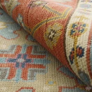 May include: Close-up of a rolled-up, patterned rug. The rug features a cream-colored base with geometric designs in shades of coral, light blue, and gray. The rolled edge reveals a detailed border with floral motifs in blue, yellow, and pink.