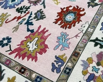 May include: A detailed close-up of a hand-knotted rug with a light pink field and a border. The rug features intricate floral and geometric patterns in shades of red, blue, green, and yellow. The border is a neutral gray.