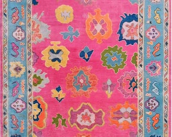May include: A vibrant pink rug with a turquoise border. The rug features a repeating pattern of colorful, abstract floral and geometric shapes in shades of yellow, orange, blue, and green. The border has a detailed design.