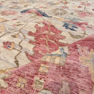 Puede incluir: Primer plano de una alfombra beige con un estampado floral en tonos rojos, rosas, azules y amarillos. La alfombra tiene un aspecto desgastado, con una sección doblada que revela la parte inferior.
