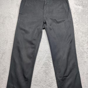 Puede incluir: Un pantalón gris oscuro con cierre de botón y bolsillos laterales. El pantalón está hecho de una tela lisa y tiene un diseño clásico de pierna recta. El pantalón está tendido sobre una superficie gris claro.