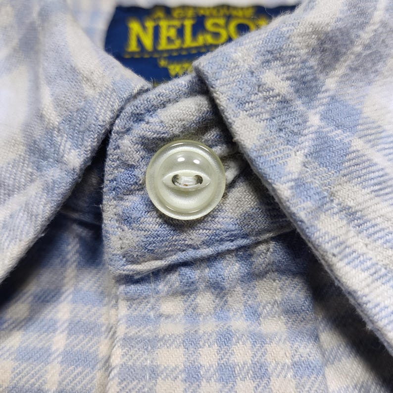 May include: Close-up of a light blue and white plaid button-down shirt. The shirt features a clear, round button and a collar. The tag reads "NELSON".