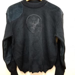 May include: A black long-sleeve sweater featuring a ribbed crew neckline, suede shoulder patches, and suede elbow patches. The chest displays a circular embroidered emblem with a deer head graphic and the text "SOUP TEAM" in black thread.