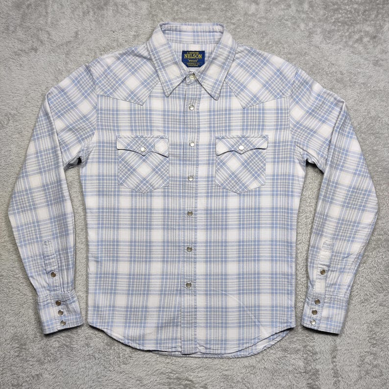 May include: A light blue and white plaid button-down shirt with long sleeves. The shirt has two chest pockets and a pointed collar. The brand name "NELSON" is visible on a small tag near the collar.