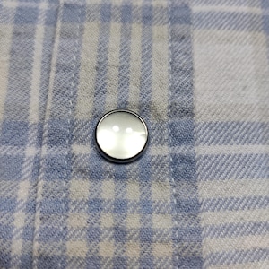 May include: Close-up of a round, pearlescent button with a silver rim, sewn onto a blue and white plaid fabric. The button has two small holes and reflects light. The fabric has a woven texture.