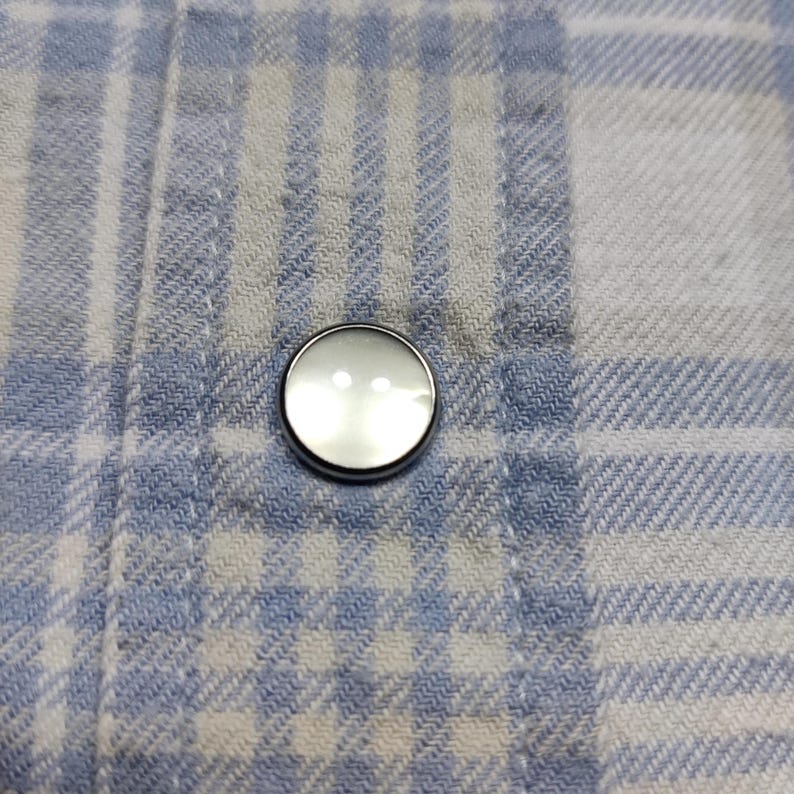 May include: Close-up of a round, pearlescent button with a silver rim, sewn onto a blue and white plaid fabric. The button has two small holes and reflects light. The fabric has a woven texture.