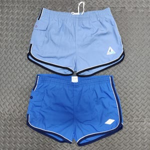 Puede incluir: Dos pares de pantalones cortos deportivos en azul claro y azul. Ambos tienen cintura con cordón, bolsillos laterales y ribetes blancos y negros. El pantalón corto azul claro tiene un logotipo triangular, el azul un logotipo de diamante.
