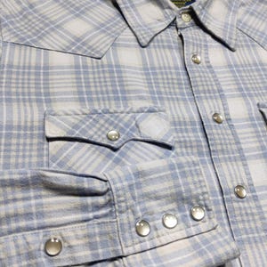 May include: A light blue and white plaid shirt with a button-down collar, two chest pockets, and snap closures. The long-sleeved shirt has snap closures on the cuffs and a Western-style yoke.