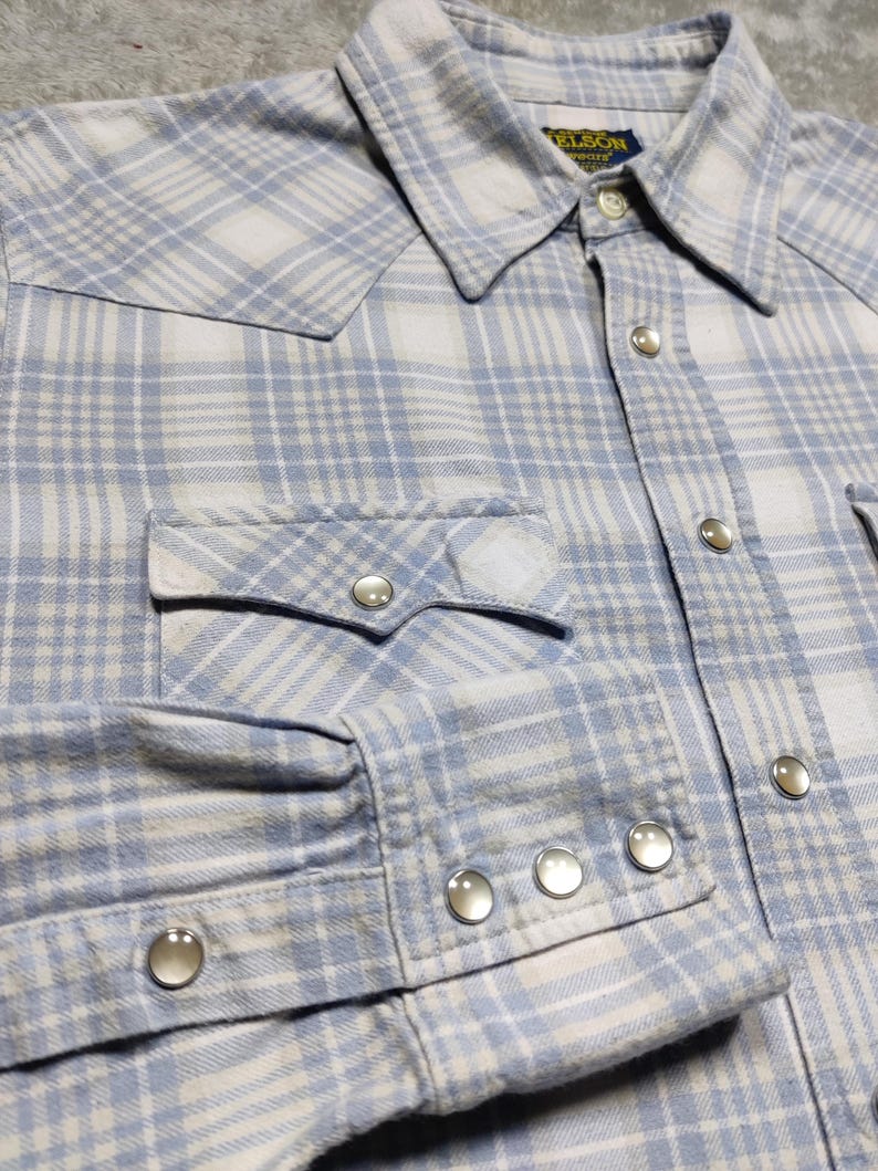 May include: A light blue and white plaid shirt with a button-down collar, two chest pockets, and snap closures. The long-sleeved shirt has snap closures on the cuffs and a Western-style yoke.