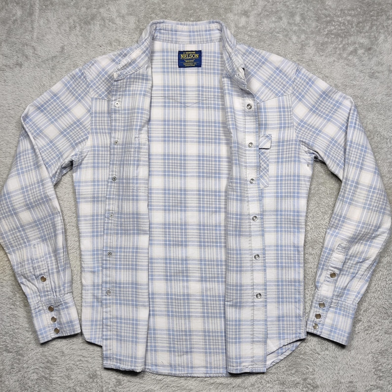 May include: A light blue and white plaid button-down shirt with long sleeves. The shirt has a classic collar, snap closures, and a small label that reads "NELSON". The shirt is open, displaying the interior.
