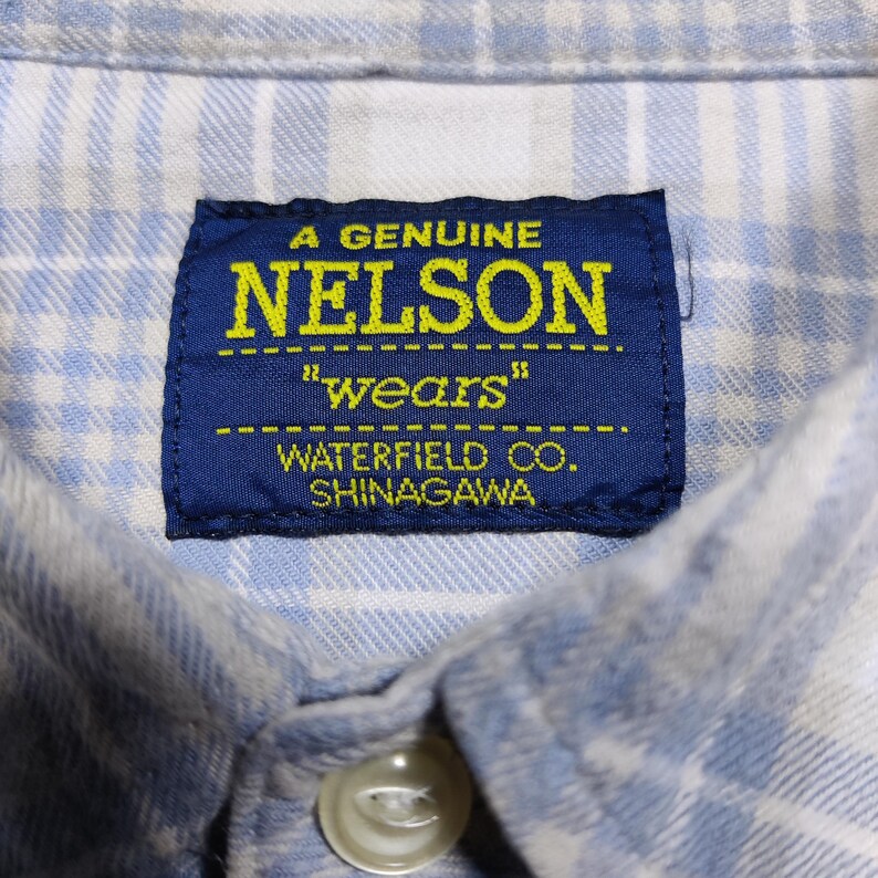 May include: Close-up of a blue and white plaid shirt collar with a button. A navy blue tag reads "A GENUINE NELSON" in yellow text, with ""wears"" below. The tag also says "WATERFIELD CO. SHINAGAWA."