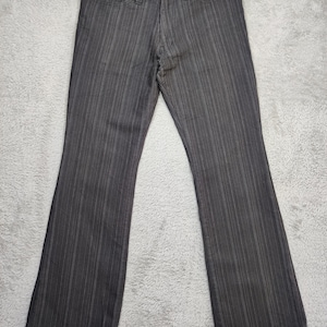 Puede incluir: Pantalones a rayas finas de color gris oscuro con pernera acampanada. Los pantalones tienen cierre de botón y cremallera, con remaches en los bolsillos. La tela tiene rayas verticales. Los pantalones están planos sobre una superficie gris.