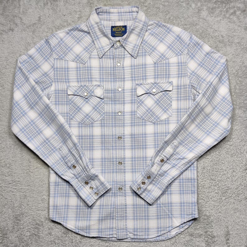 May include: A light blue and white plaid button-down shirt with long sleeves. The shirt has two chest pockets and a collar. The brand name "NELSON" is on a small tag at the collar.