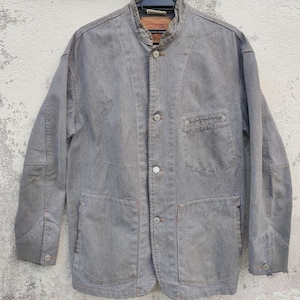 Puede incluir: Chaqueta vaquera gris con cuello mao, cierre de botones y múltiples bolsillos. La chaqueta tiene un aspecto descolorido y un pequeño bolsillo en el pecho. Una etiqueta roja y blanca es visible cerca del cuello.