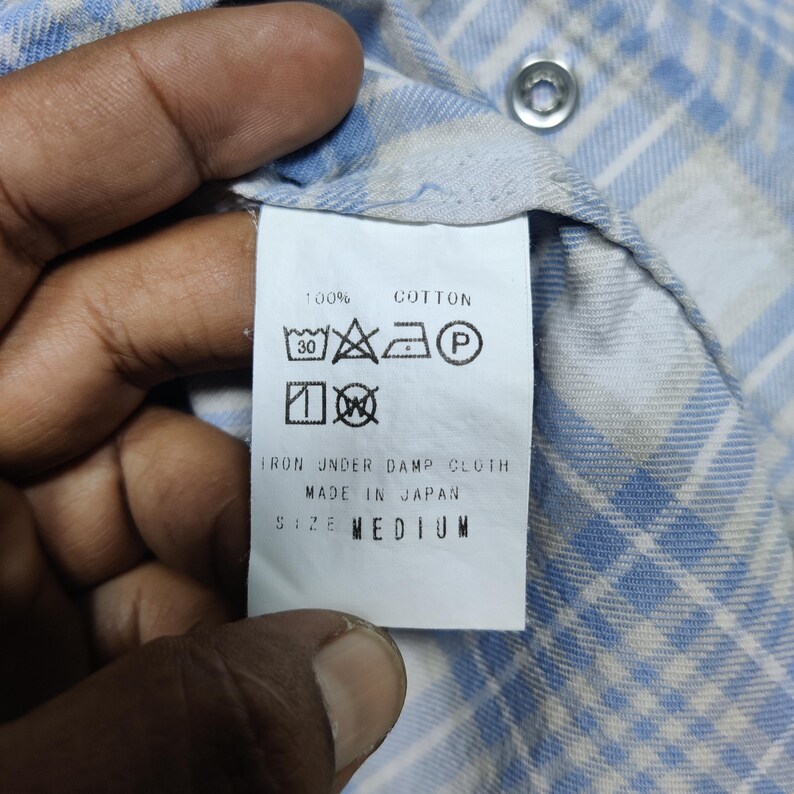 May include: Close-up of a white tag attached to a blue and white plaid shirt. The tag reads "100% Cotton" and includes care symbols. The text "IRON UNDER DAMP CLOTH" and "MADE IN JAPAN" are also visible. The size is "MEDIUM".