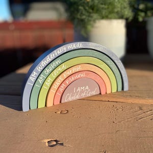 May include: A wooden rainbow stacker in pastel colors with inspirational phrases. The rainbow features layers of blue, green, yellow, orange, and pink, each inscribed with positive affirmations like "I am loved" and "Child of God."