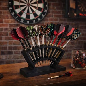 May include: A black dart holder with a selection of darts, showcasing vibrant flights, sits on a wooden surface. A pint of beer and a dartboard are in the background. A jar of darts and a copper mug are also visible.