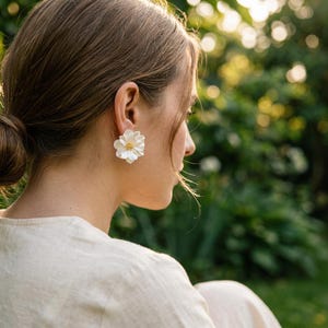 Op de afbeelding: Witte bloemvormige oorstekers met een gouden centrum. De oorbellen zijn het middelpunt van de afbeelding, tegen een vage groene achtergrond. De oorbellen hebben een delicaat bloemenontwerp.