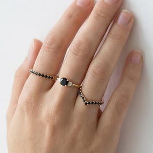 May include: A hand displays three gold rings. One ring features a row of small black stones, another has a large black stone with two smaller white stones, and the third is a V-shaped ring with black stones.