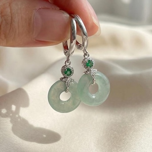 Könnte beinhalten: Silberne Creolen-Ohrringe mit Jadeit-Donut-Anhängern. Jeder Ohrring hat einen kleinen grünen Edelstein und silberne Filigranarbeiten. Der Jadeit hat eine hellgrüne Farbe. Die Ohrringe werden von einer Hand gehalten.