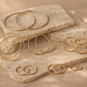 May include: A collection of gold-colored hoop earrings of varying sizes, arranged on a textured, light-colored stone surface. The earrings are simple, circular designs, reflecting light. The image showcases several pairs, highlighting the different diameters available.