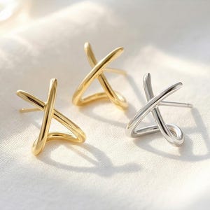 May include: Three pairs of stud earrings in gold and silver tones. Each earring features an abstract X-shaped design. The earrings are displayed on a white, textured surface, with soft lighting and shadows.