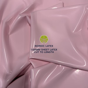 May include: A sheet of light pink latex with a glossy, reflective surface. The material is folded, creating soft drapes and shadows. Printed in the center is a green circular logo with the text NORDIC LATEX 1.00MM SHEET LATEX CUT TO LENGTH.