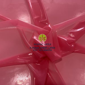 May include: A sheet of pink latex with a glossy, reflective surface. The material is folded to create depth and highlights. Printed on the centre is a green circular logo with the text NORDIC LATEX and 0.40MM SHEET LATEX CUT TO LENGTH.