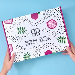 Two hands holding the signature pomegranate-patterned Balm Box, showcasing the box design and pink edges against a light blue background.
