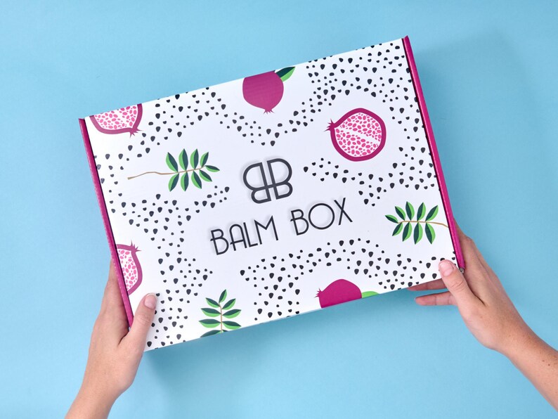 Two hands holding the signature pomegranate-patterned Balm Box, showcasing the box design and pink edges against a light blue background.