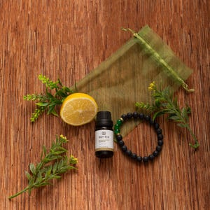 Op de afbeelding: Een wellness-thema stilleven met een halve citroen, een klein flesje etherische olie, een zwarte kralenarmband met een groene accent en een groene mesh zak. Op een houten oppervlak met takjes groen.