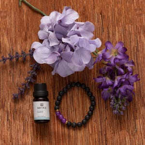 Könnte beinhalten: Eine schwarze Flasche ätherisches Öl, ein schwarzes Perlenarmband mit lila Akzenten und lila Blumen sind auf einer Holzoberfläche angeordnet. Die Ölflasche hat ein weißes Etikett. Das Armband hat einen goldenen Akzent.