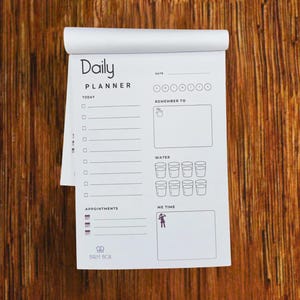 May include: A white daily planner notepad with the words "Daily PLANNER" at the top. The notepad has sections for "Today", "Appointments", "Remember To", "Water", and "Me Time". The notepad is on a wooden surface.