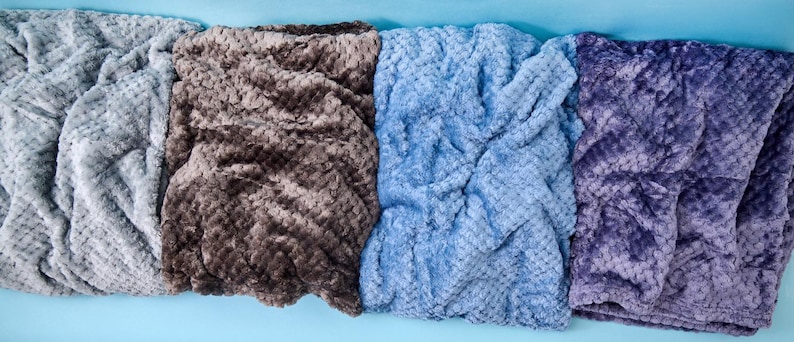 Soft plush fuzzy blankets in Dove Grey, Campfire, Ash Blue, and Lilac folded side by side, Balm Box cozy throw