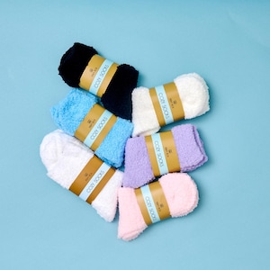 May include: A collection of six pairs of fuzzy socks in black, white, light blue, lavender, and light pink. Each pair is rolled and secured with a tan paper band featuring the text COZY SOCKS and a small logo printed in black ink.