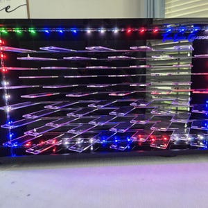 May include: A rectangular black display case with multiple clear acrylic shelves arranged in a grid. The unit features integrated LED lighting strips along the sides that emit red, blue, and green light. The text True Laser Creations is visible.