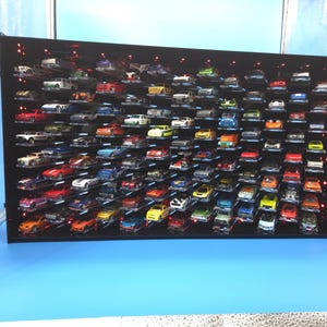 May include: A black rectangular wall-mounted display case featuring multiple horizontal shelves filled with numerous small die-cast toy cars in various colors. The unit includes integrated red LED lighting along the interior edges.