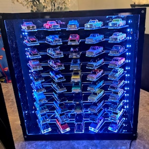 May include: A black rectangular display case with a clear front panel, illuminated by internal blue LED lights. The case features multiple horizontal shelves holding a collection of small die-cast toy trucks in various colors and designs.