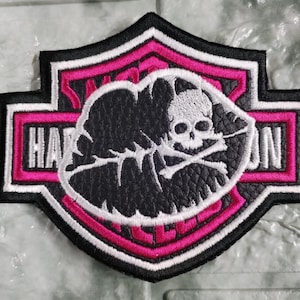 Puede incluir: Un parche bordado en forma de escudo con un borde negro y rosa que incluye el texto Harley-Davidson en blanco. El centro muestra un óvalo negro con textura de cuero y un gráfico bordado en blanco de una calavera con huesos cruzados.