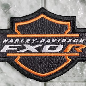 May include: A black and orange embroidered patch featuring a shield-shaped design with a textured leather-like appearance. The text Harley-Davidson is embroidered in white, with the letters FXDR in white and orange across the center of the shield.