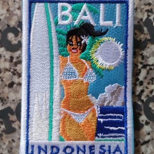 Può includere: Una toppa rettangolare ricamata con la scritta BALI in alto e INDONESIA in basso. Il disegno raffigura una persona in bikini bianco accanto a una tavola da surf bianca alta, con un sole luminoso, foglie di palma verdi e onde del mare blu.