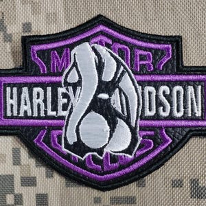 Może przedstawiać: Wyszywana naszywka w kształcie tarczy z czarno-fioletowym logo Harley-Davidson i białym napisem. Centralny srebrno-czarny element graficzny nakłada się na tekst Harley-Davidson, całość wykończona jest fioletowymi obwódkami.