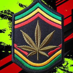 May include: A shield-shaped embroidered patch featuring a central olive-green cannabis leaf design on a black background, framed by concentric chevron stripes in vibrant green, yellow, and red colors with a thick black stitched border.