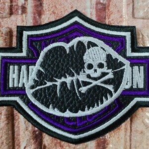 May include: A shield-shaped embroidered patch featuring a black and purple border with white accents. The centre displays a black textured oval containing a white skull and crossbones graphic. The text HARLEY-DAVIDSON is partially visible in white.