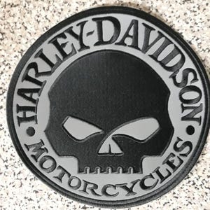 Op de afbeelding: Een ronde patch met een zwarte schedelafbeelding gecentreerd op een grijze achtergrond. De tekst HARLEY-DAVIDSON MOTORCYCLES is in zwarte letters langs de buitenrand van de cirkel aangebracht en omcirkelt het centrale schedelontwerp.