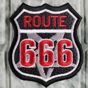 Pode incluir: Um emblema em forma de escudo com textura de couro sintético preto. A seção superior apresenta o texto bordado em vermelho ROUTE, enquanto a seção inferior exibe o grande número 666 bordado em vermelho sobre um design de pentagrama cinza.