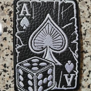 Pode incluir: Um emblema bordado retangular com fundo de couro sintético preto e um design de carta de ás de espadas bordado em branco. O design inclui um grande símbolo de espadas, a letra A nos cantos e um dado branco em primeiro plano.